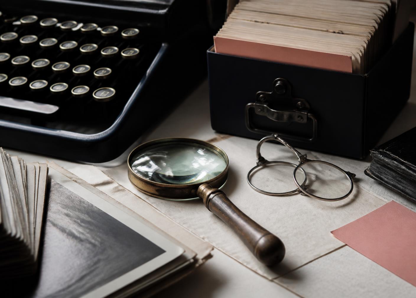 A vintage typewriter, archival index cards, magnifying glass and reading glasses on a research desk