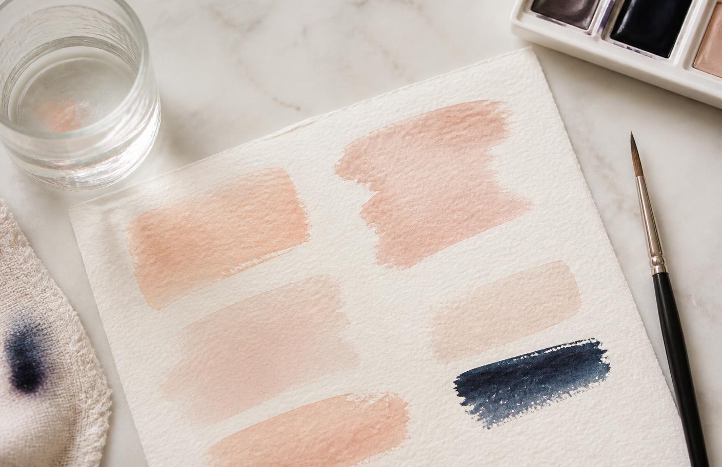 Soft peach watercolour swatches drying on textured cream paper beside a brush